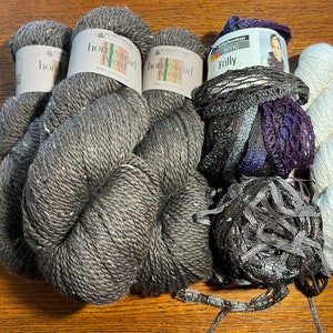 Op de afbeelding: Drie bollen donkergrijs Homespun Worsted garen, één bol wit garen en één bol zwart en paars garen met een label waarop "Frilly" staat.