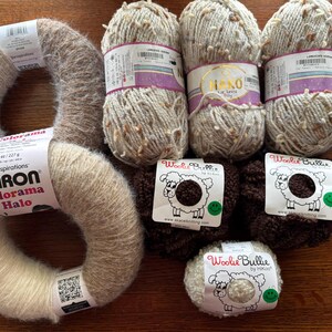 Op de afbeelding: Een verzameling garenklossen in verschillende tinten beige en bruin, waaronder een klos Caron Colorama Halo garen. De klossen zijn gelabeld met de merknamen "Woolie Bullie" en "Caron".