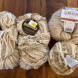 Op de afbeelding: Vier bollen garen in verschillende tinten beige en wit. Het garen is gerangschikt op een houten oppervlak. Het garen is geëtiketteerd met "Noro" en "Galway Roving".