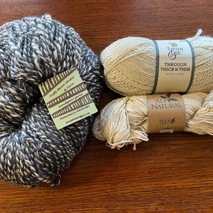 Op de afbeelding: Vier bollen garen in verschillende kleuren en texturen. De bollen liggen op een houten oppervlak. Het garen is gelabeld met "Tuffy Barn Studio & Farm", "Yarn Bee Through Thick & Thin" en "Alp Natural Feza".