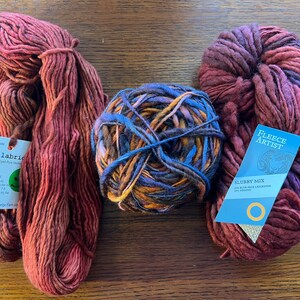 Op de afbeelding: Drie bollen garen in verschillende kleuren en texturen. De bol links is diep rood en oranje van kleur met een label waarop "Malabrigo Kettle Dyed Fine Merino" staat en informatie over het garen bevat. De bol in het midden is een mix van blauw, paars, oranje en bruin. De bol rechts is diep rood en paars van kleur met een label waarop "Fleece Artist Slubby Mix 50% Blue Faced Leicester 50% Merino" staat.
