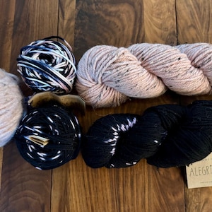 Op de afbeelding: Vier bollen garen in verschillende kleuren en patronen. Twee bollen zijn roze met zwarte vlekken, één is zwart met witte vlekken en één is zwart met roze vlekken. Het garen is geëtiketteerd met "Woolstok Tweed" en "Alegria Grande".