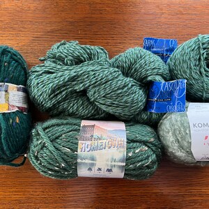 Op de afbeelding: Vijf bollen groene garen. Het garen is in verschillende texturen en tinten groen. De garenetiketten bevatten de merknamen "Lopi", "Cascade Yarns" en "Komorebi".