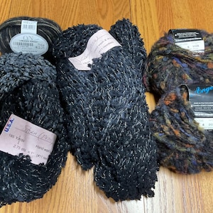 Op de afbeelding: Drie bollen garen in verschillende kleuren en texturen. De eerste bol is zwart met zilveren spikkels en een label met de tekst "Stitch (Price) 185 Yards 2.75 ozs. 1 - (11 mm) Hand wash". De tweede bol is zwart met zilveren spikkels. De derde bol is een mix van bruin, zwart en blauw met een label met de tekst "Longoo 2 (Dark) Street Charles, Inc. 6 0120".
