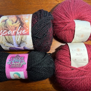Op de afbeelding: Vier bollen garen in verschillende kleuren en merken. Het garen is rood, zwart en een mix van rood en zwart. De etiketten op het garen zeggen "scarfie", "Rowan Big Wool" en "I Love This Chunky".