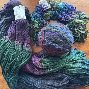 Op de afbeelding: Een assortiment kleurrijk garen in tinten groen, paars en blauw. Het garen is gerangschikt op een houten oppervlak, met een bolletje veelkleurig garen en een bundel lintachtig materiaal. Een label is bevestigd aan een van de garenbundels.