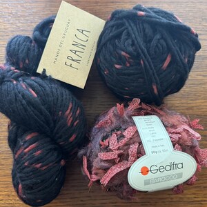 Op de afbeelding: Drie strengen zwart garen met rode spikkels en een bolletje getextureerd, veelkleurig garen. Eén streng heeft een label met de tekst "FRANCA" en "MANOS DEL URUGUAY". De andere bol garen heeft een label met de tekst "Gedifra Barocco".