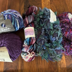 Op de afbeelding: Assortiment van kleurrijke garen strengen in paars, blauw en teal. Inclusief een bol paars garen, een streng met het label "Flamy Lala Berlin" en een streng met het label "Ridiculous Hand Dyed Yarn". Perfect voor brei- en haakprojecten.