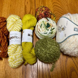 Op de afbeelding: Zes bollen garen in verschillende kleuren en texturen, waaronder bruin, geel, groen en wit. Het garen is in een rij op een houten oppervlak gerangschikt.