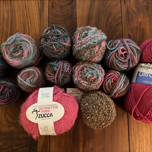 Op de afbeelding: Een verzameling garenbollen in verschillende kleuren en texturen, waaronder roze, bruin, blauw en groen. De garenbollen zijn gerangschikt op een houten oppervlak. Een garenbol heeft een label met de tekst "Trendsetter Yarns Zucca Made in Italy".