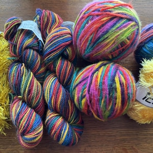 Op de afbeelding: Een assortiment van kleurrijke garenklossen en bollen, waaronder een gele, pluche garenbol met het label "Ice Yarns".