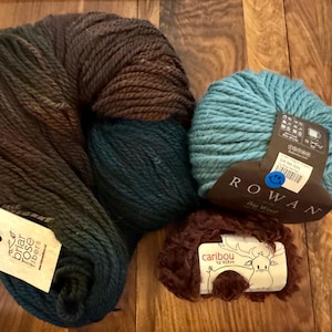 May include: Three skeins of yarn in different colors and textures. The first skein is a dark brown and teal yarn with a label that says "Briar Rose Fibers". The second skein is a light blue yarn with a label that says "Rowan". The third skein is a brown fuzzy yarn with a label that says "caribou by Eileen".