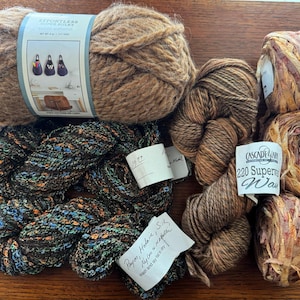Op de afbeelding: Een verzameling verschillende garenbollen in bruin, beige en veelkleurige tinten. De afbeelding toont een grote bol bruin garen met een label, verschillende kleinere bollen en een lintgaren. Ideaal voor brei- en haakprojecten.
