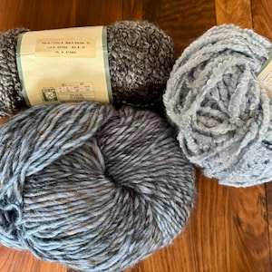 Op de afbeelding: Drie bollen garen in verschillende kleuren en texturen. De bol links is donkergrijs met een gevlekt patroon. De bol in het midden is een blauw-grijze mix met een grove textuur. De bol rechts is lichtblauw met een pluche textuur. Het etiket op de bol links luidt "Special Color: Black Marble 102 Lot # 837024 Art # 87 WL # 612463".