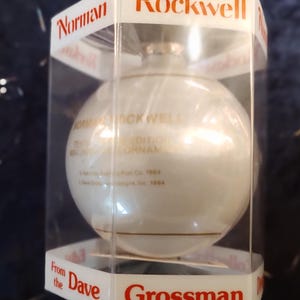 May include: A white glass Christmas ornament with gold lettering that reads "Norman Rockwell Edition Ornament". The ornament is in a clear plastic box with a red label that reads "Rockwell" and "Grossman".