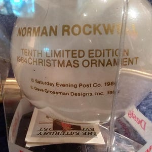 May include: A white glass Christmas ornament with gold lettering that reads "NORMAN ROCKWELL TENTH LIMITED EDITION 1984 CHRISTMAS ORNAMENT &copy; Saturday Evening Post Co. 1984 Dave Grossman Designs, Inc. 1984".