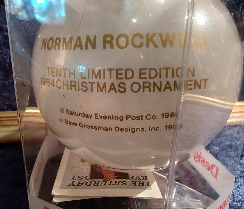 May include: A white glass Christmas ornament with gold lettering that reads "NORMAN ROCKWELL TENTH LIMITED EDITION 1984 CHRISTMAS ORNAMENT &copy; Saturday Evening Post Co. 1984 Dave Grossman Designs, Inc. 1984".