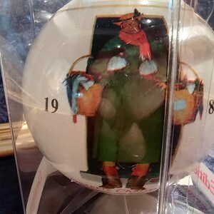 May include: A white glass Christmas ornament with a painted image of a person carrying a basket. The ornament is numbered 19 and 84.