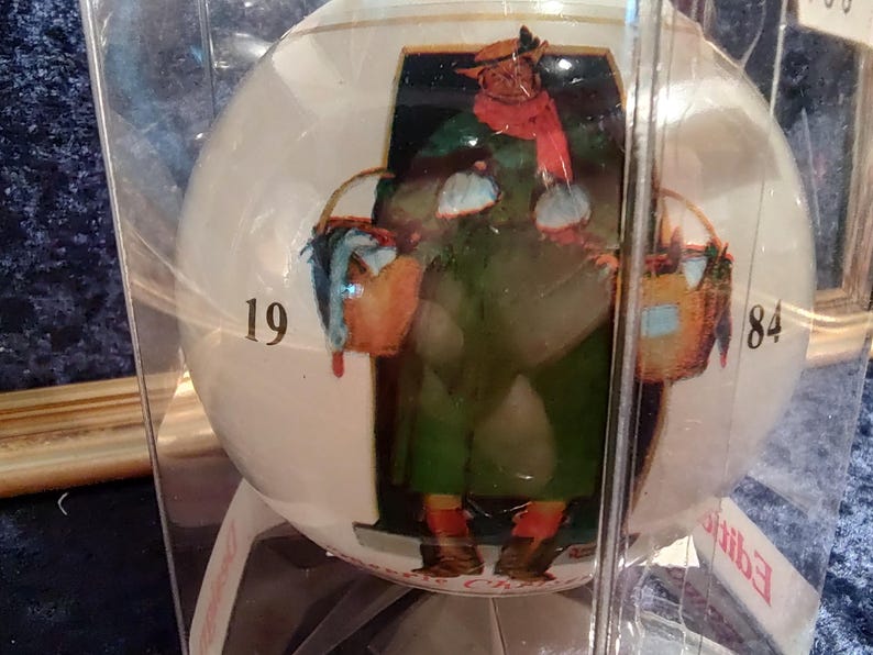 May include: A white glass Christmas ornament with a painted image of a person carrying a basket. The ornament is numbered 19 and 84.