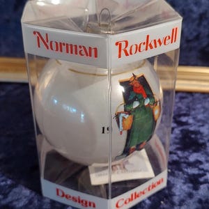 May include: A white glass Christmas ornament with a Norman Rockwell design featuring a woman carrying a basket. The ornament is in a clear plastic box with the words "Norman Rockwell Design Collection" printed on the bottom.