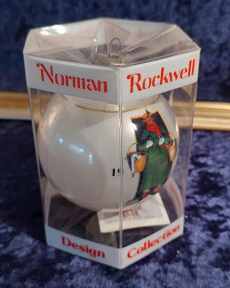May include: A white glass Christmas ornament with a Norman Rockwell design featuring a woman carrying a basket. The ornament is in a clear plastic box with the words "Norman Rockwell Design Collection" printed on the bottom.