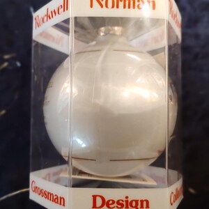 May include: A white glass Christmas ornament with gold trim in a clear plastic box. The box has the words "Norman Rockwell Grossman Design Collection" printed on it.