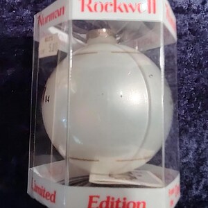 May include: A white glass Christmas ornament with a gold trim, in a clear plastic box. The box has the words "Rockwell" and "Limited Edition" printed on it.