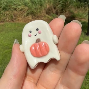 May include: A small, white, glossy ceramic ghost figurine featuring a smiling face with pink cheeks and a small orange pumpkin attached to its center. The ghost has rounded arms extending to the sides and a smooth, curved bottom edge.