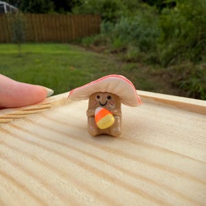 May include: A whimsical, handmade mushroom figurine with a smiling face and a candy corn. The mushroom has a light brown stem and a pink and white cap. It is holding a candy corn. The figurine is on a wooden surface.