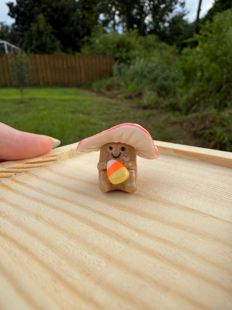May include: A whimsical, handmade mushroom figurine with a smiling face and a candy corn. The mushroom has a light brown stem and a pink and white cap. It is holding a candy corn. The figurine is on a wooden surface.