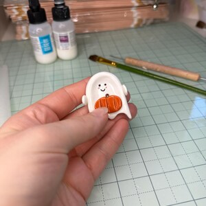 May include: A small, white, ghost-shaped ceramic or clay charm featuring a smiling face with black eyes and pink cheeks. An orange, textured pumpkin is attached to the center of the ghost's body, which includes a small hole for threading.