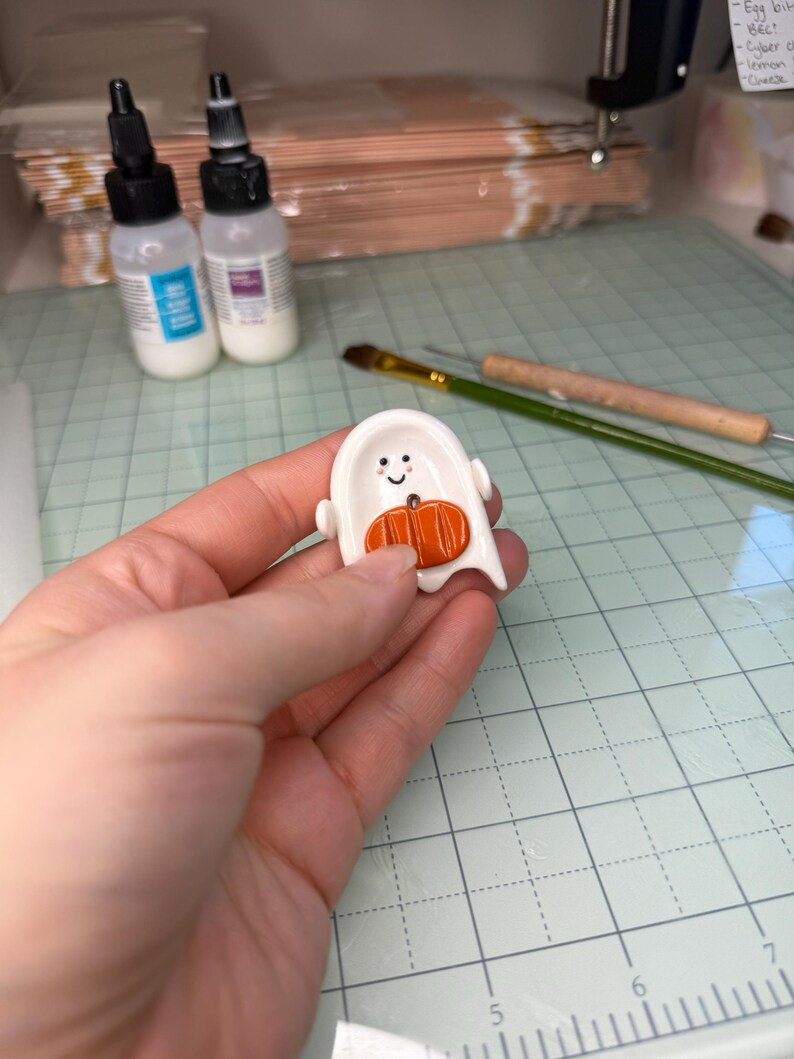 May include: A small, white, ghost-shaped ceramic or clay charm featuring a smiling face with black eyes and pink cheeks. An orange, textured pumpkin is attached to the center of the ghost's body, which includes a small hole for threading.