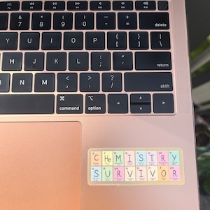 May include: A rose gold laptop with a sticker on the trackpad. The sticker is a periodic table with the word "CHEMISTRY" spelled out using the element symbols. Below the word "CHEMISTRY" is the word "SURVIVOR" spelled out using the element symbols.