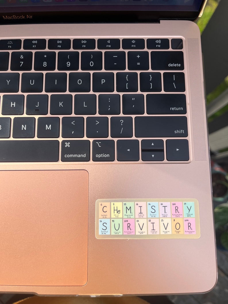 May include: A rose gold laptop with a sticker on the trackpad. The sticker is a periodic table with the word "CHEMISTRY" spelled out using the element symbols. Below the word "CHEMISTRY" is the word "SURVIVOR" spelled out using the element symbols.