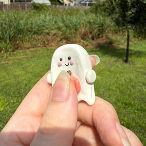 May include: A small white ghost figurine crafted from clay, featuring two black dot eyes, two pink circular cheeks, a small curved black mouth, and a protruding orange tongue. The ghost has rounded side arms and a smooth, rounded top surface.