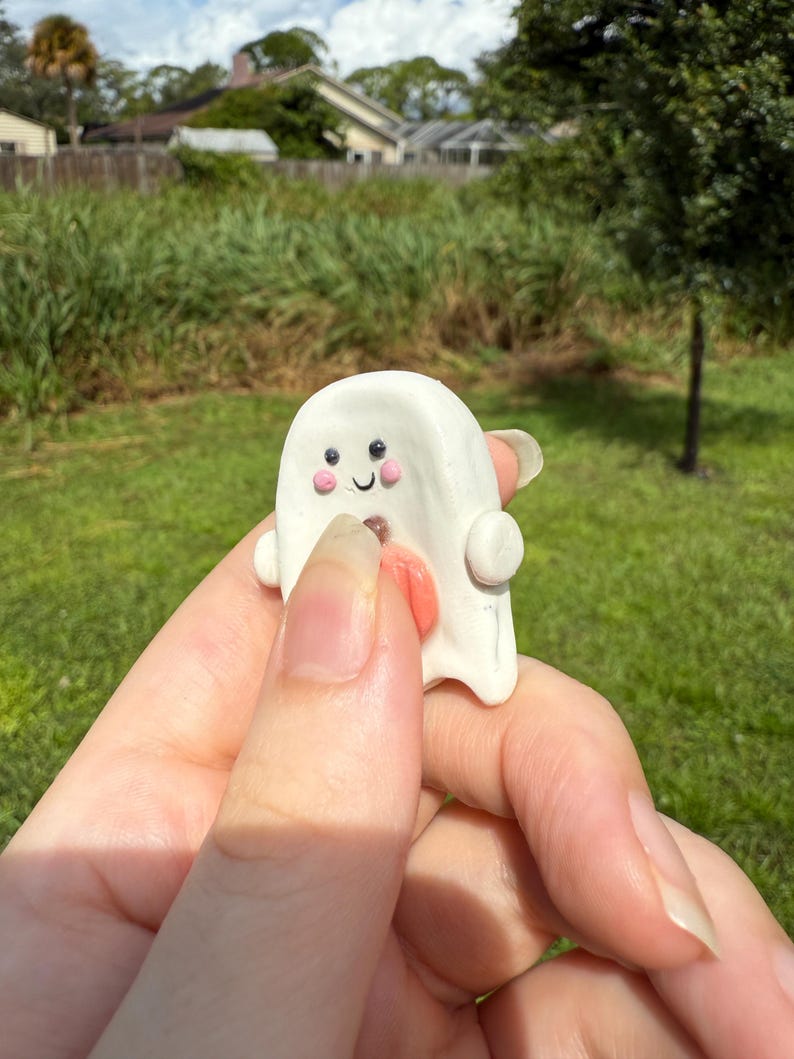 May include: A small white ghost figurine crafted from clay, featuring two black dot eyes, two pink circular cheeks, a small curved black mouth, and a protruding orange tongue. The ghost has rounded side arms and a smooth, rounded top surface.