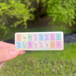 May include: A sticker with the word "CHEMISTRY" spelled out using the chemical symbols for the elements. Below the word is the word "SURVIVOR" spelled out using the chemical symbols for the elements. The sticker is being held by a hand.