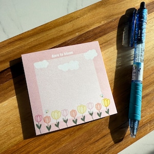 May include: A pink notepad with the text "Born to bloom" and a floral border of tulips. A blue and clear pen rests on a wooden surface next to the notepad.