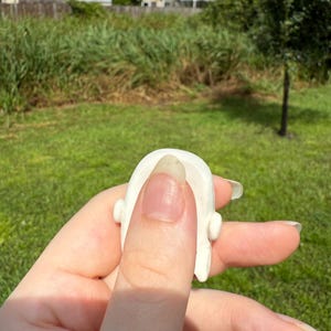 May include: A small, smooth, white ceramic object with a rounded top and two small protruding side elements. The piece has a curved, arch-like shape and a solid, opaque finish, appearing as a singular, compact decorative component.