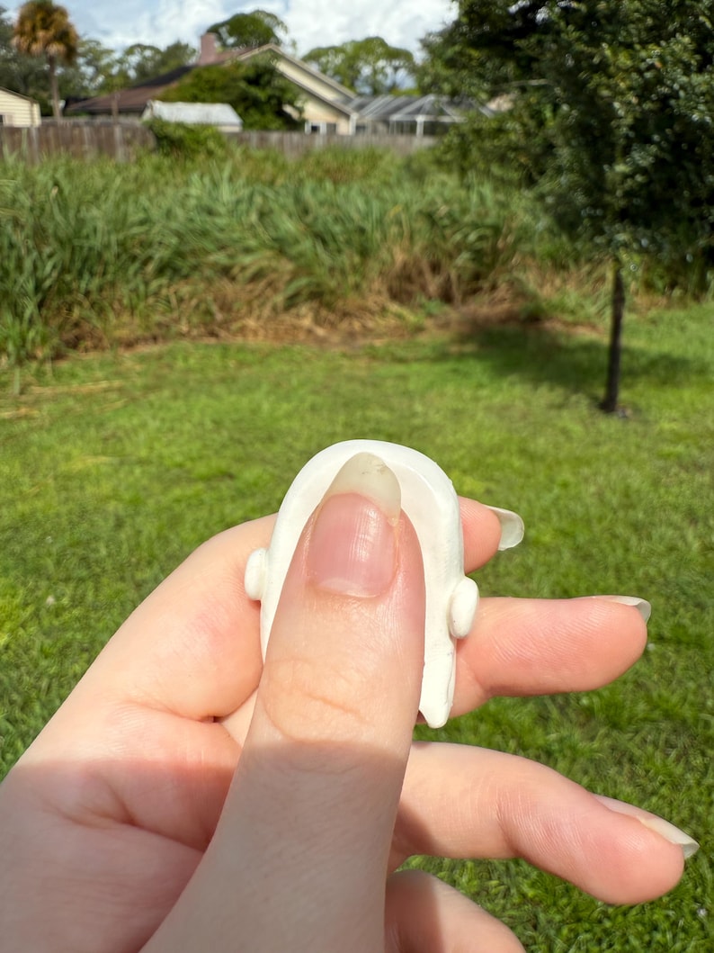 May include: A small, smooth, white ceramic object with a rounded top and two small protruding side elements. The piece has a curved, arch-like shape and a solid, opaque finish, appearing as a singular, compact decorative component.