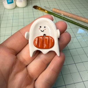 May include: A small, white, ghost-shaped figurine made of smooth material, featuring a painted black smiling face with pink cheeks and a raised, glossy orange pumpkin attached to the center of its body. The ghost has two small arms extended outward.