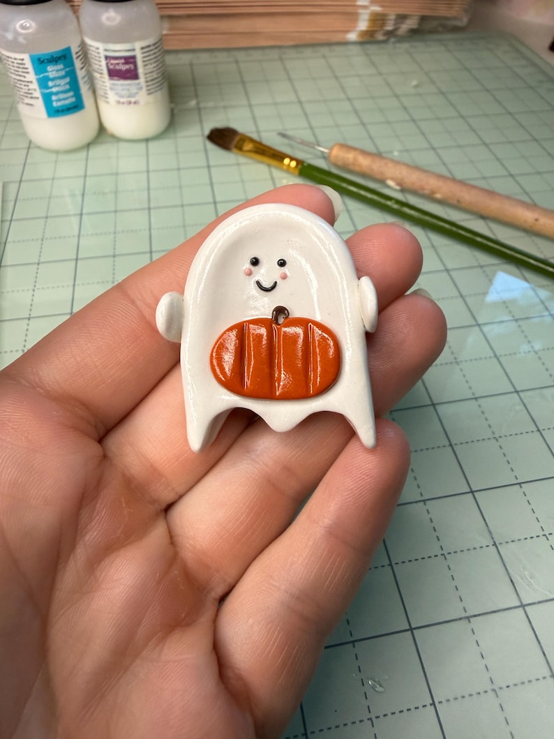 May include: A small, white, ghost-shaped figurine made of smooth material, featuring a painted black smiling face with pink cheeks and a raised, glossy orange pumpkin attached to the center of its body. The ghost has two small arms extended outward.