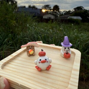 May include: A wooden tray holds three whimsical clay figurines. A mushroom with a candy corn, a white chick with a pumpkin, and a ghost with a pumpkin. The figurines are in a natural outdoor setting with a sunset in the background.