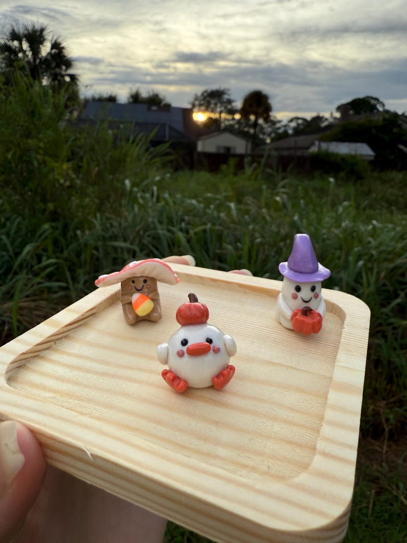 May include: A wooden tray holds three whimsical clay figurines. A mushroom with a candy corn, a white chick with a pumpkin, and a ghost with a pumpkin. The figurines are in a natural outdoor setting with a sunset in the background.
