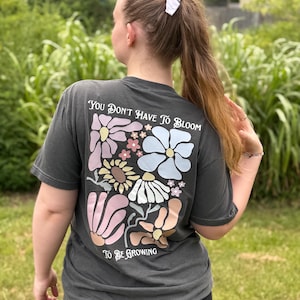 Puede incluir: Camiseta gris oscuro con un diseño floral en la espalda. El diseño incluye varias flores en colores pastel y el texto "You Don't Have To Bloom To Be Growing". Una prenda de vestir informal.