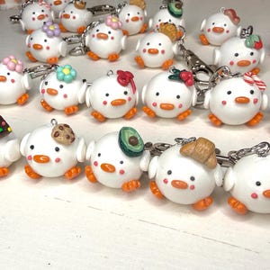 May include: A collection of white chick-shaped keychains with orange feet and beaks. Each keychain is decorated with a different miniature food item or accessory, such as flowers, bows, or a cookie. The keychains are attached to silver metal rings.