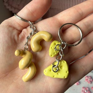 May include: Two keychains with food-themed charms. One keychain features two yellow macaroni charms. The other keychain has a yellow cheese wedge charm with holes. Both keychains have silver-colored metal rings and chains.
