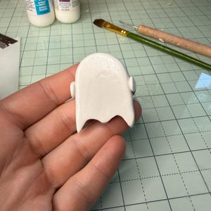 May include: A smooth, white, rounded clay figure with a scalloped bottom edge and small side protrusions. The object has a glossy finish and is held in a palm, with craft supplies including two bottles of Sculpey glaze and a brush visible nearby.