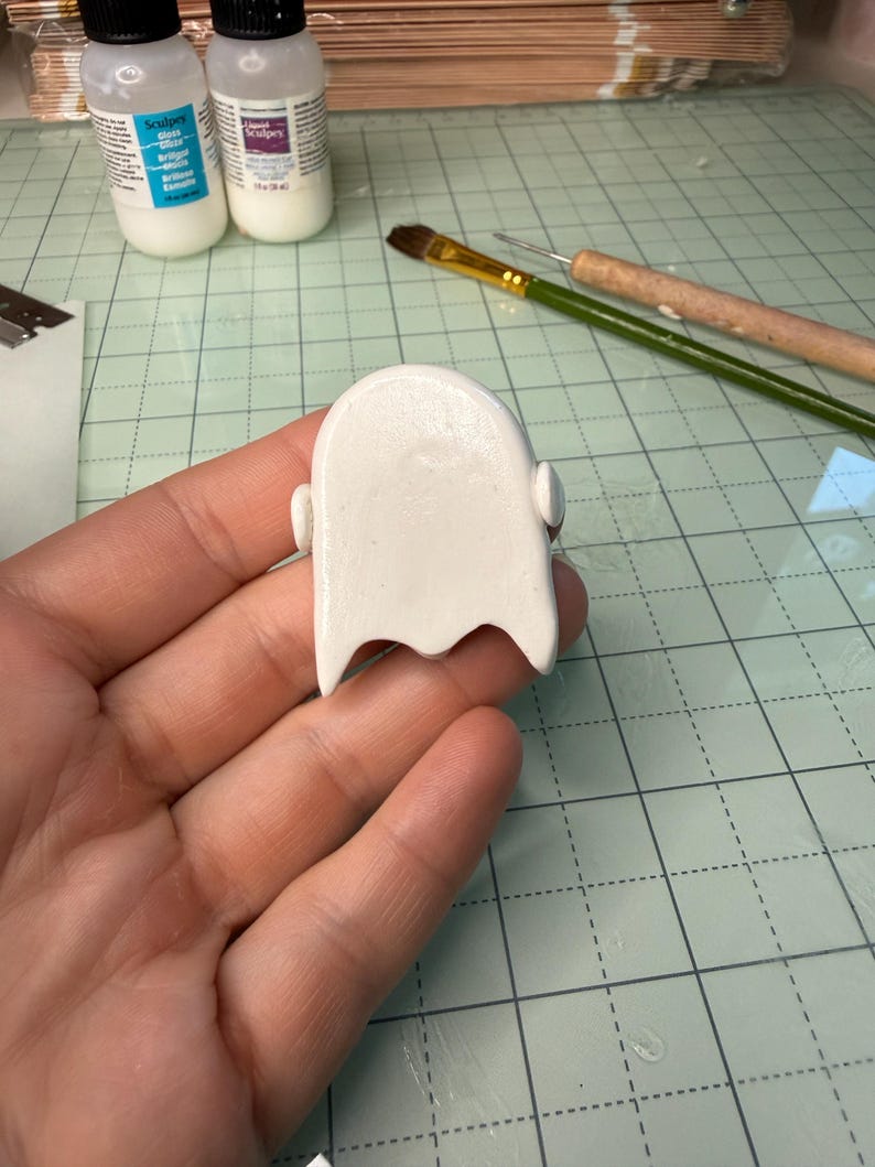 May include: A smooth, white, rounded clay figure with a scalloped bottom edge and small side protrusions. The object has a glossy finish and is held in a palm, with craft supplies including two bottles of Sculpey glaze and a brush visible nearby.