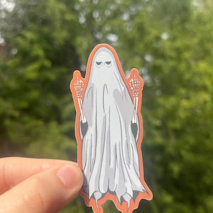 May include: A white ghost with a grumpy expression is giving the middle finger with both hands. The ghost's hands are skeletal and the sticker has an orange outline.